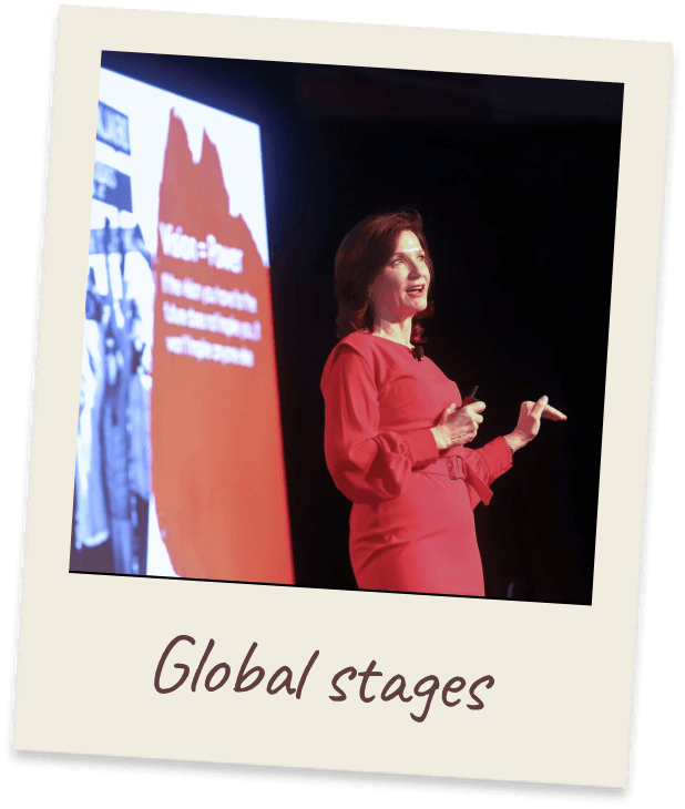 Global stages. Margie in a red dress speaking on a dark stage beside a large presentation screen.