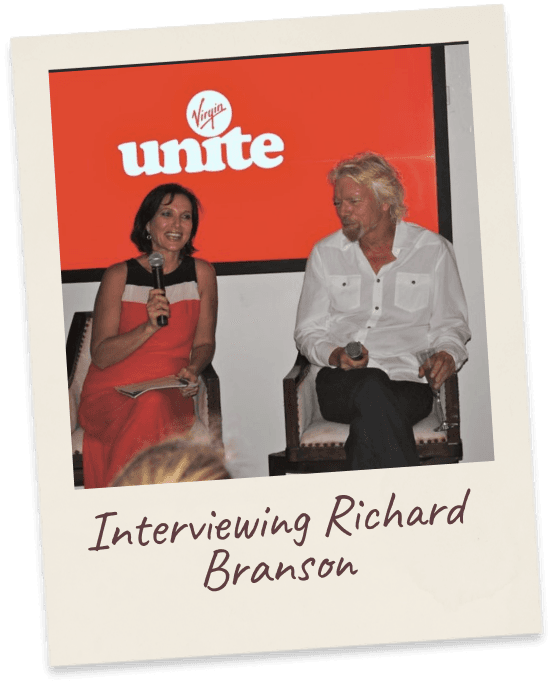 Interviewing Richard Branson. Margie in red on stage with a microphone, in conversation with Richard Branson in front of a Virgin Unite backdrop.