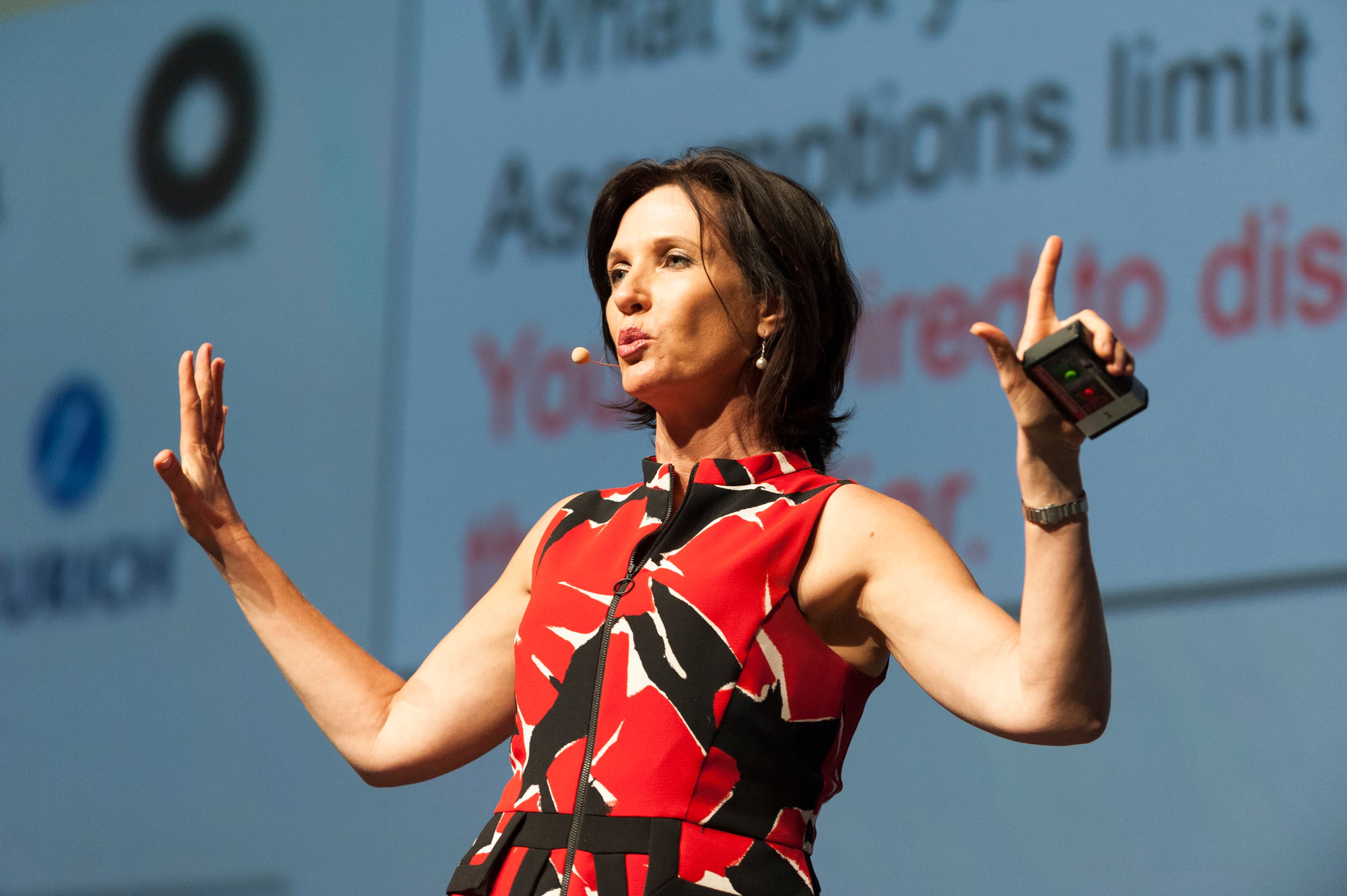 Margie Warrell mid-keynote on stage in a red and black patterned dress, arms wide, a presentation screen behind her
