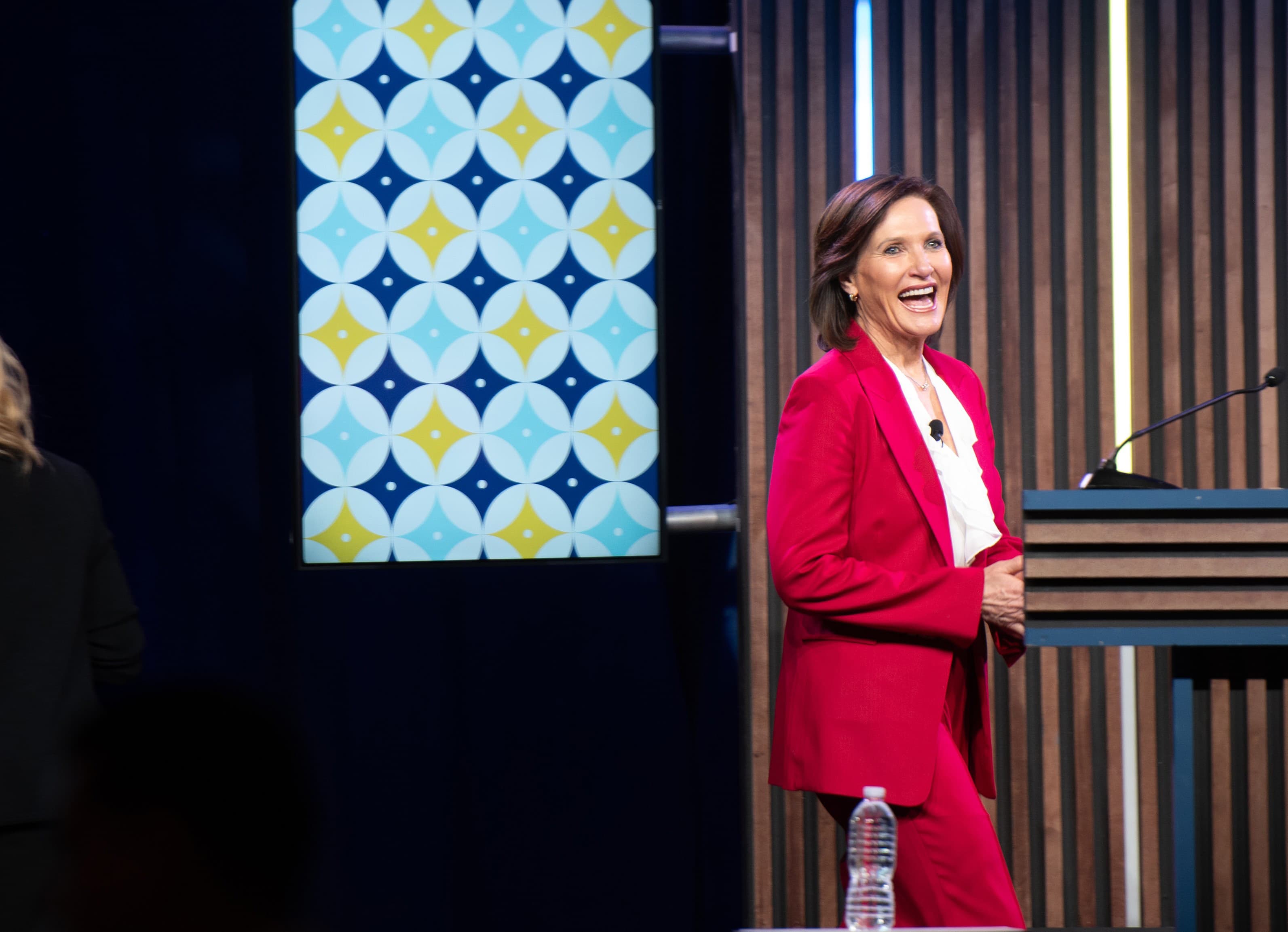Dr. Margie Warrell speaking on a dark stage in a crimson trouser suit and white blouse, laughing mid-sentence beside a timber-slat lectern, in front of a tall banner printed with a repeating circle-and-four-point-star pattern