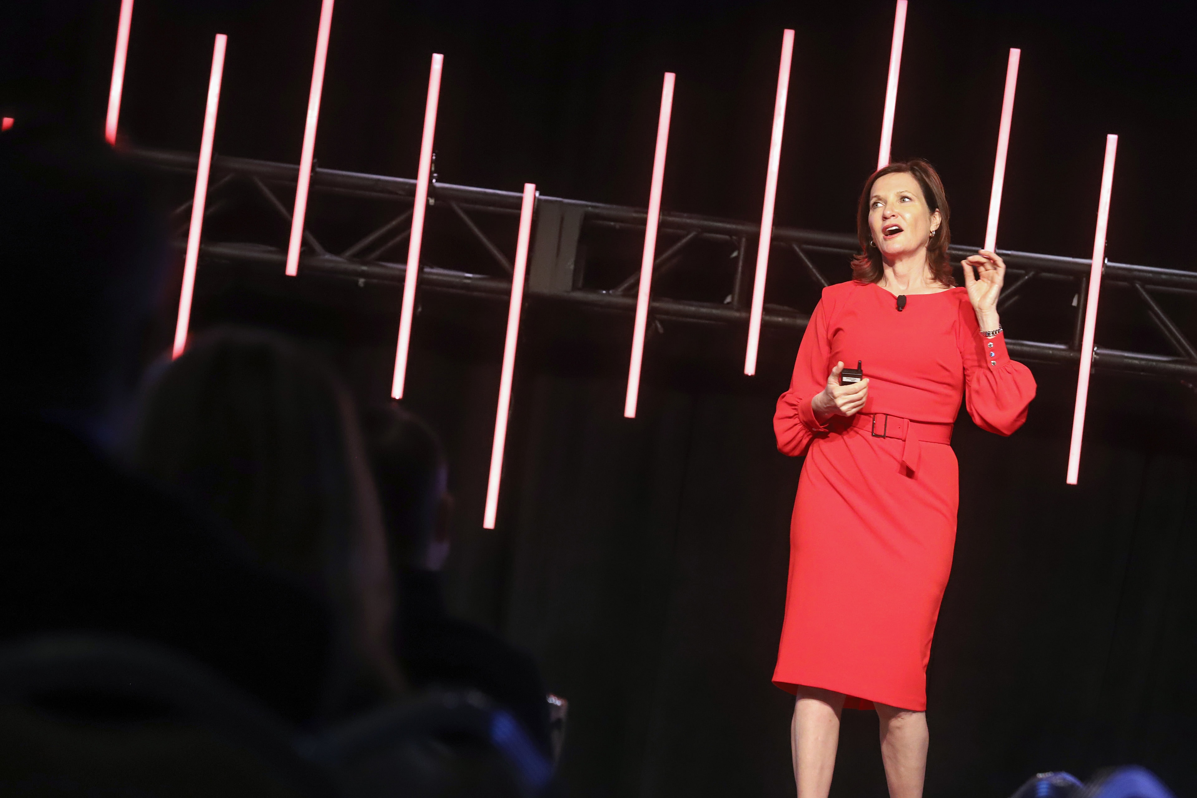 Dr. Margie Warrell on stage in a red dress, speaking beneath a truss of vertical red light bars, the audience in silhouette