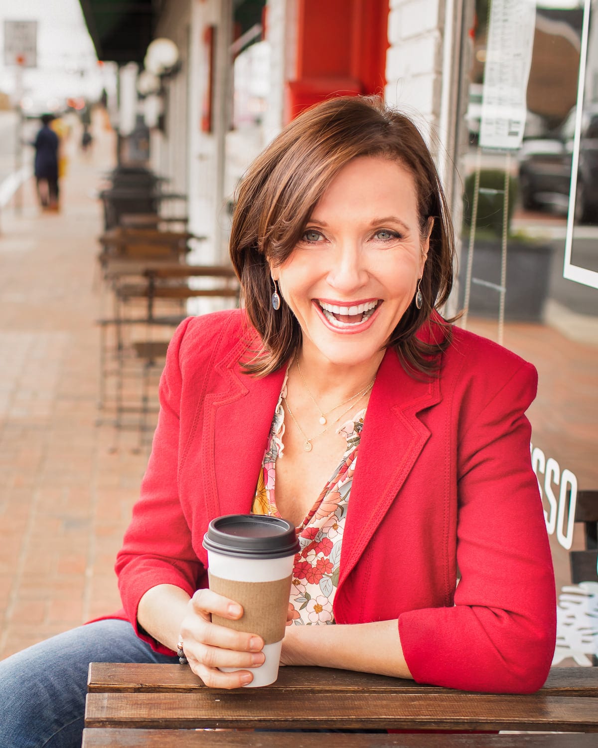 Dr. Margie Warrell laughing at a street-side café table, coffee in hand, in a red blazer