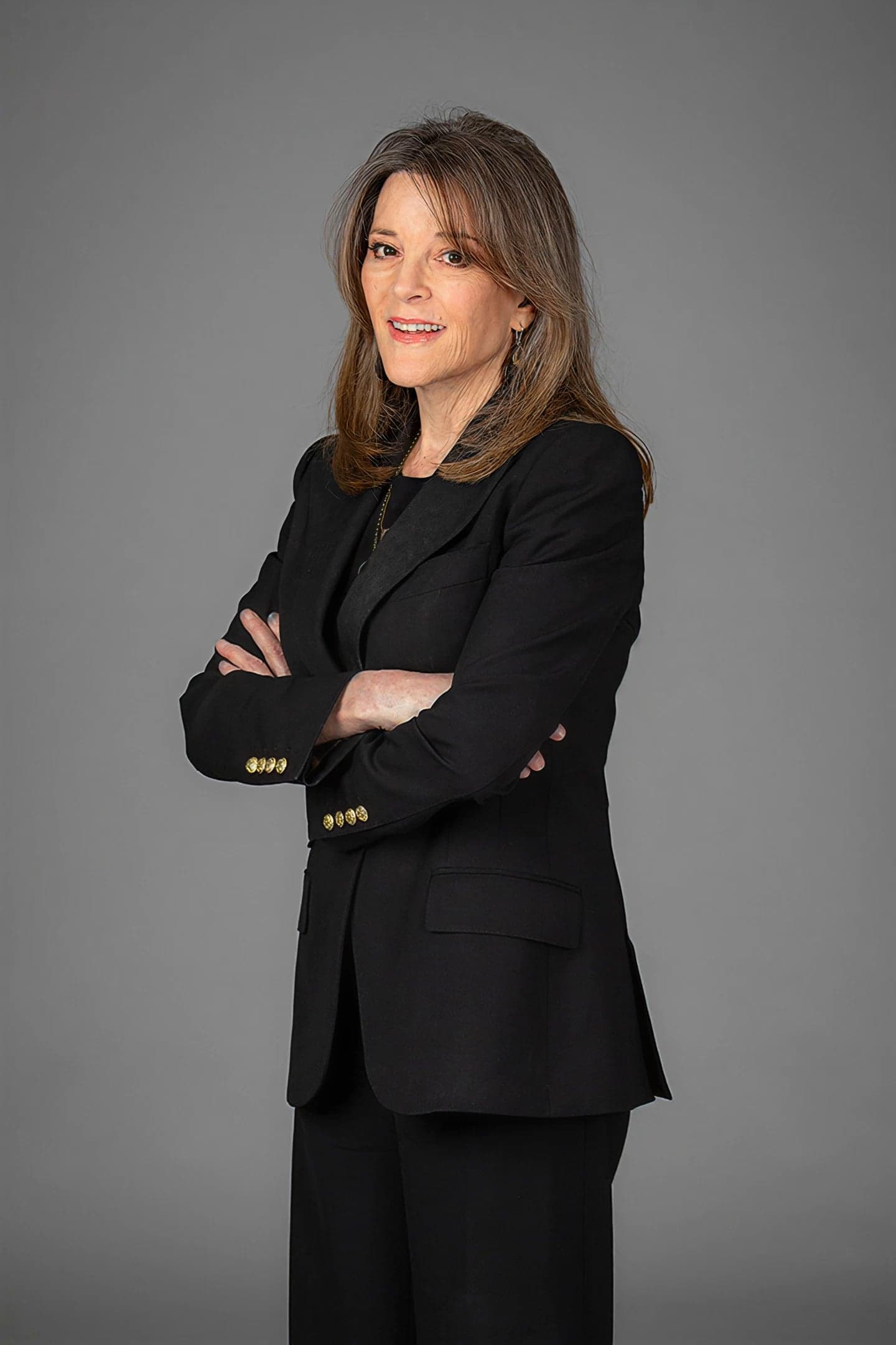 Marianne Williamson, spiritual author and presidential candidate — arms folded, in a dark jacket, against a grey studio backdrop