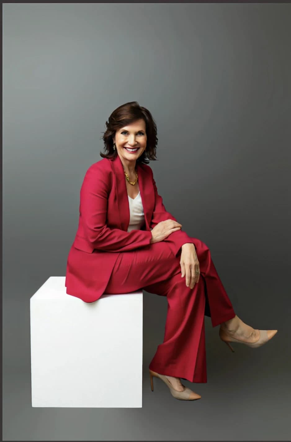 Dr. Margie Warrell full length in a crimson trouser suit over a white top with nude heels, perched on a white cube plinth with her legs extended, on a graduated mid-grey backdrop